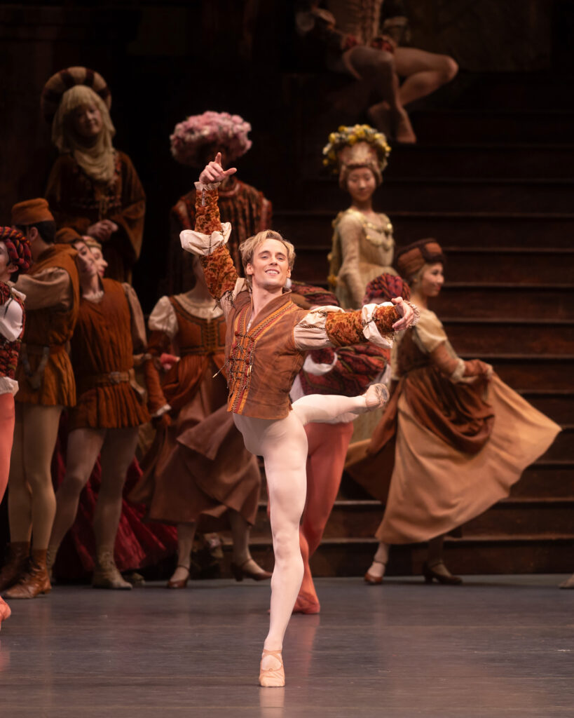 Jonathan Klein does an attitude derriére during a performance of Romeo and Juliet onstage. He wears a rust colored jacket with puffed sleeves and off-white tights and ballet slippers, and he looks out towards the audience with a joyful smile.