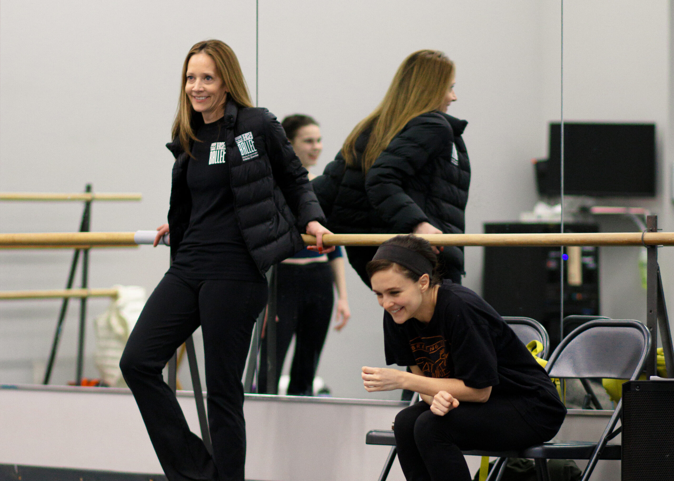 Maria Kowroski, wearing black yoga pants, a black shirt and a puffy black vest, leans against a ballet barre in a dance studio, her back facing the mirror. Next to her on her left, Lauren Lovette, in a black t-shirt and black yoga pants, sits in a gray fold-out chair, leaning forward with her forearms resting on her thighs, and smiles. A female dancer is seen in the background through the mirror.