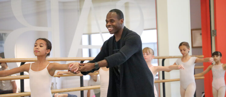 Christopher Charles McDaniel, wearing a long black cardigan sweater over a dark t-shirt and track pants, smiles and adjusts the arms of a young female ballet student during a ballet class. The children wear white leotards and hold their arms n second position.
