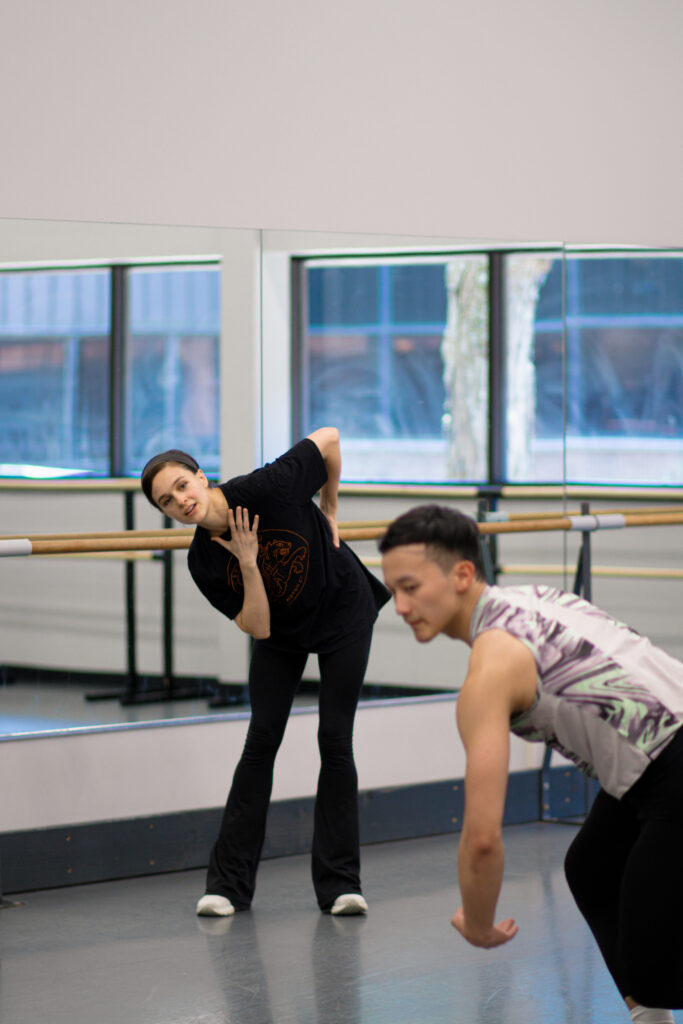 Lauren Lovette, in a black t-shirt, yoga pants and sneakers, Leans to her right as she watches a male dancer dance in a ballet studio. Her back is toward a mirror along the wall.