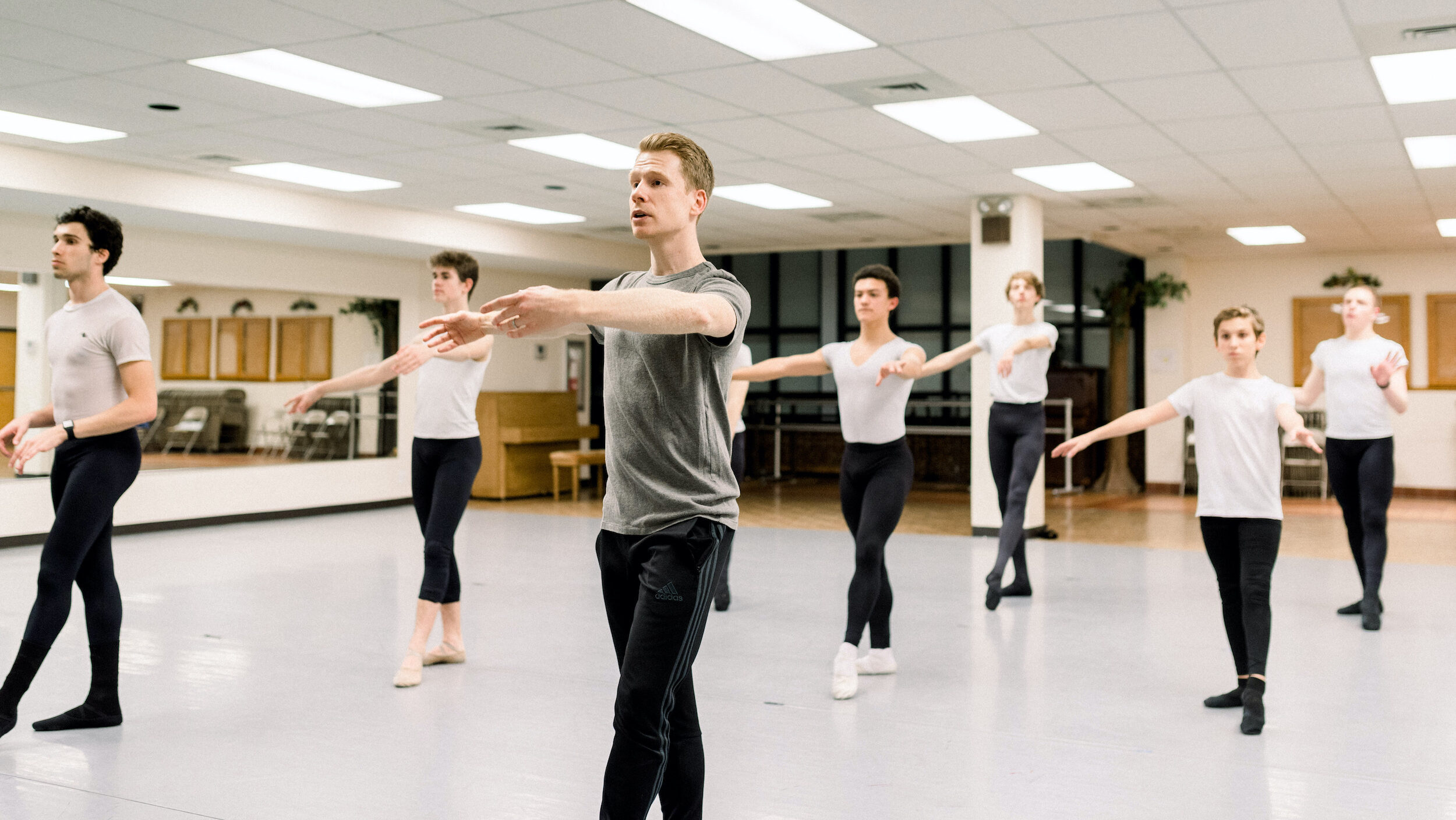A male ballet teacher stands at the front of a dance studio in tendu à la quatrieme devant with his left leg. He wears a gray t-shirt and black track pants. Behind him, seven teenage boys in white t-shirts, black tights and black ballet slippers practice the same move.