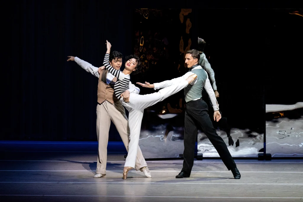 Mikaela Santos and two male dancers perform a pas de trois onstage. Santos takes an attitude derrierre position on pointe and wraps her arms around Masero's shoulders as he holds her around the waist. Morawski stands to her left in tendu back and reaches for her with his left arm. Santos wears white pants and a black and white striped long-sleeved shirt. The men wear trousers and button down vests with white shirts underneath.