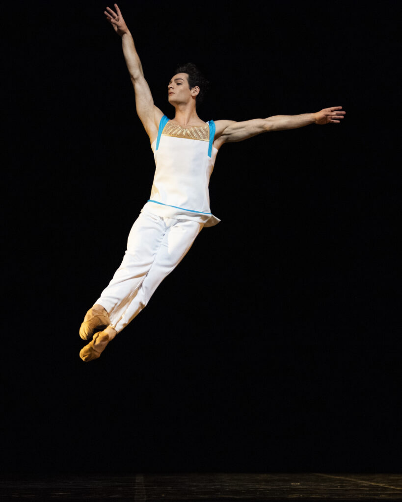 Angelo Greco flies through the air in an assemblé position. He wears white pants and a mostly white top with bright blue straps.