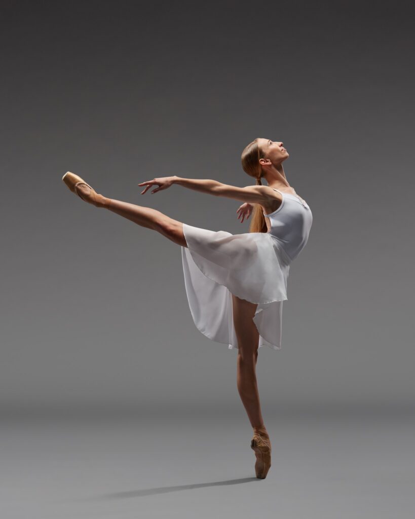 Catherine Conley, a ballerina, poses on pointe in an arabesque, pressing both arms back behind her as she looks up. She wears a white leotard and knee-length white wrap skirt. She poses in front of a gray backdrop.