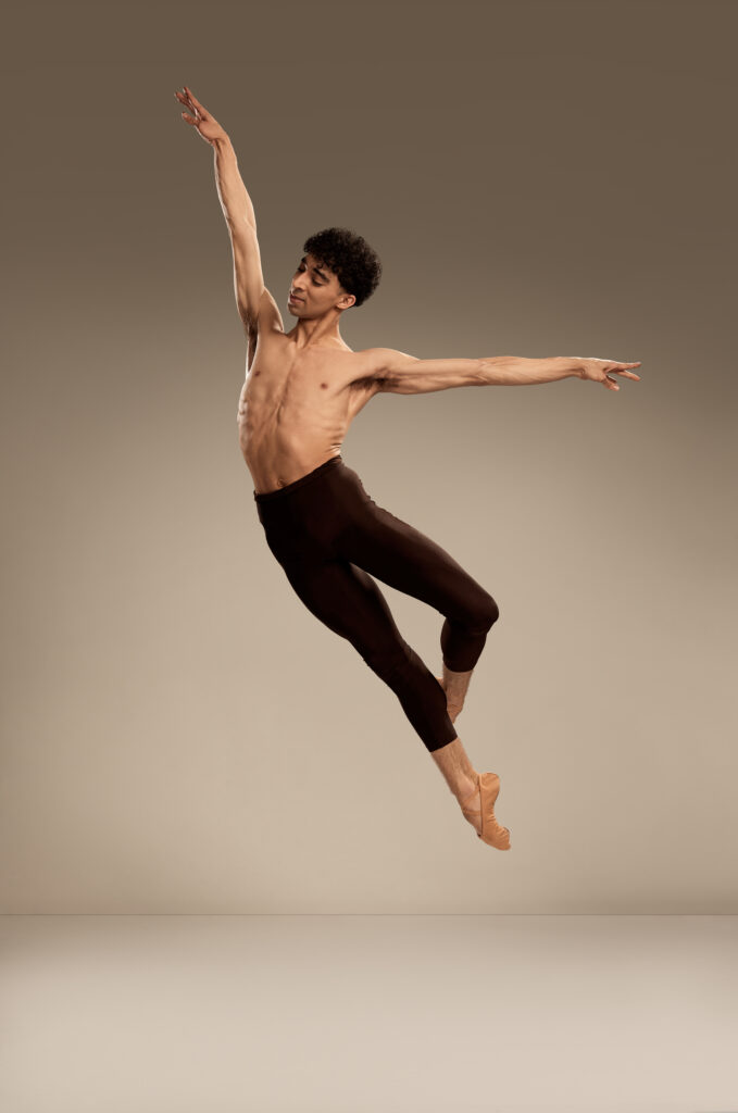 Cato Berry jumps up with his left leg in coupé back, his right arm raised, and his left arm out to the side. He is shirtless and wears dark-colored tights and tan ballet slippers. He jumps in front of a white backdrop.