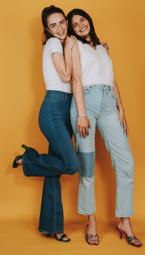 ELise Kruger and Sophie Milosevic, two women in their 20s or early 30s, stand close to each other in front of a yellow backdrop. They both wear jeans, white t-shirts and high heels. Kruger leans against Milosevic and puts both hands on her shoulders.