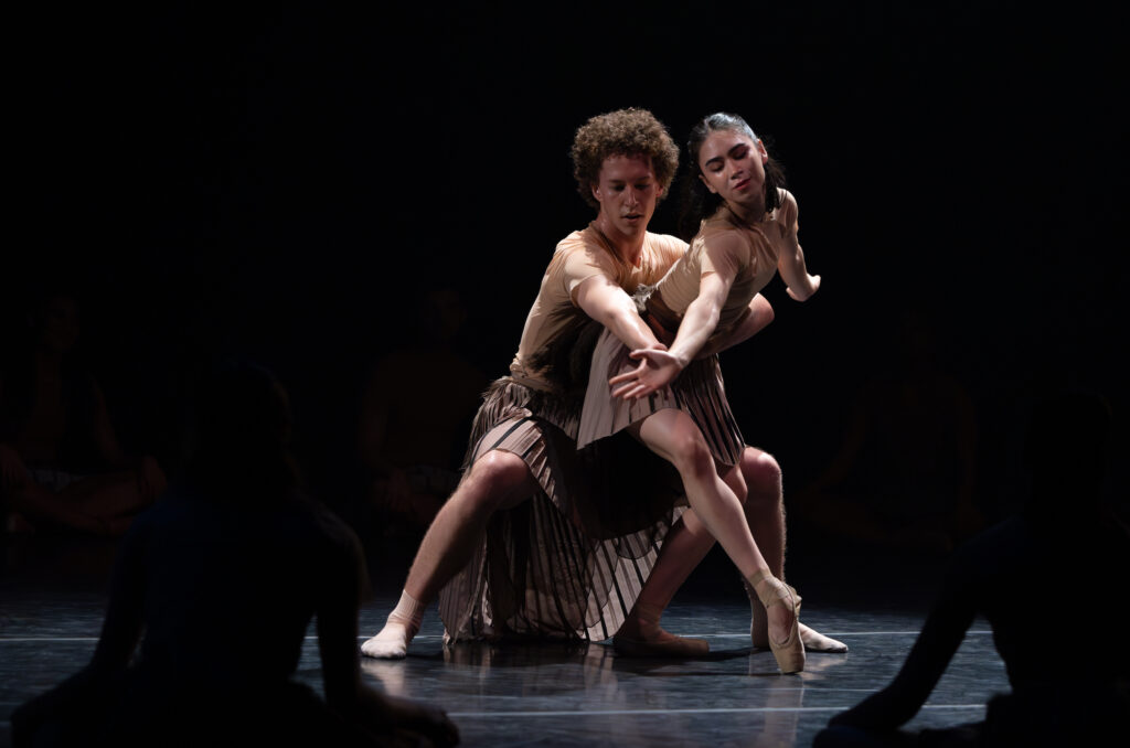 During a performance onstage, dancer Luke Csordas squats slightly in second position and holds Evelyn Robinson be the waist with his left arm. She reaches forward with her right hand as Csordas touches her wrist with his right hand. She pliés and props her right foot up onto pointe. They both wear brown pleated skirts and mesh tops.