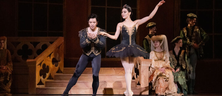 During a performance of Swan Lake, Evelyn Robinson, in a black tutu with gold trim and a gold tiara, stands in sus-sous on pointe. She playfully pulls her right hand away from her partner, Oliver Oguma, as he leans slightly forward to kiss it. He wears a black tunic with gold trim and black tights and ballet slippers.