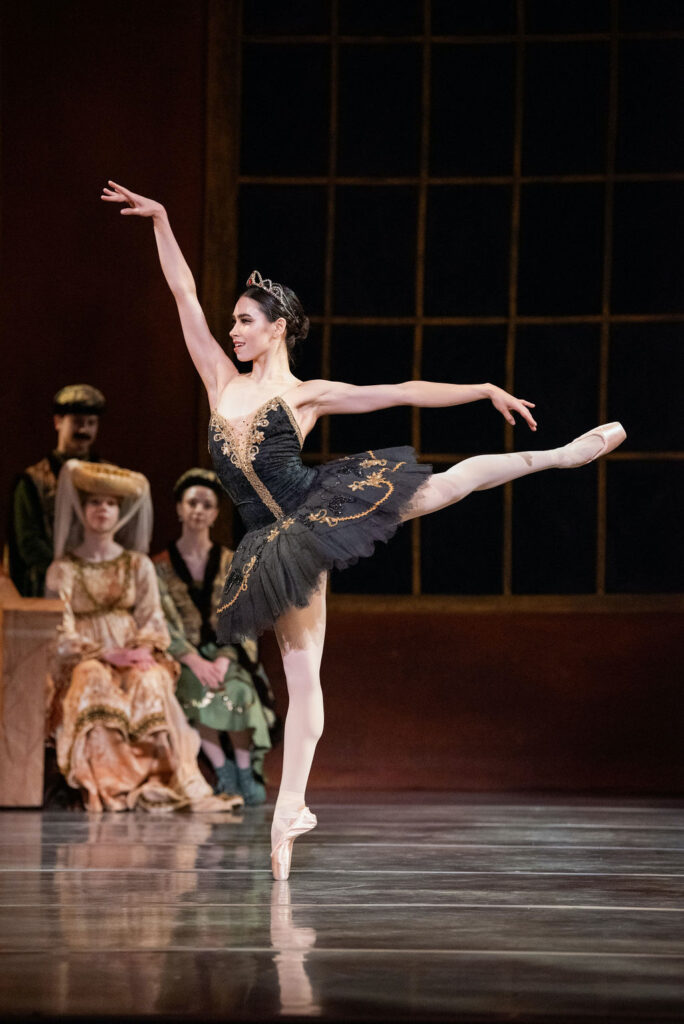 During a performance of Swan Lake, Evelyn Robinson does a first arabesque on pointe towards stage right with her left leg raised. She wears a black tutu with gold trim, and gold tiara, pink tights and pointe shoes. Dancers dressed a nobles sit in the background and watch.