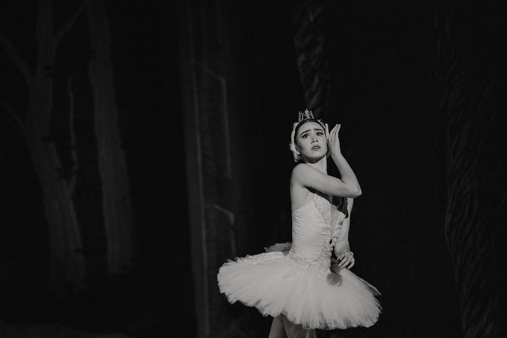 During a performance of Swan lake, Evelyn Robinson is shown from the thighs up in a white tutu with feathered trim and a feathered headpiece and crown. She looks out, her eyes filled with fear, and touches the left side of her face with the back of her right hand as she pulls her left arm in close to her torso.