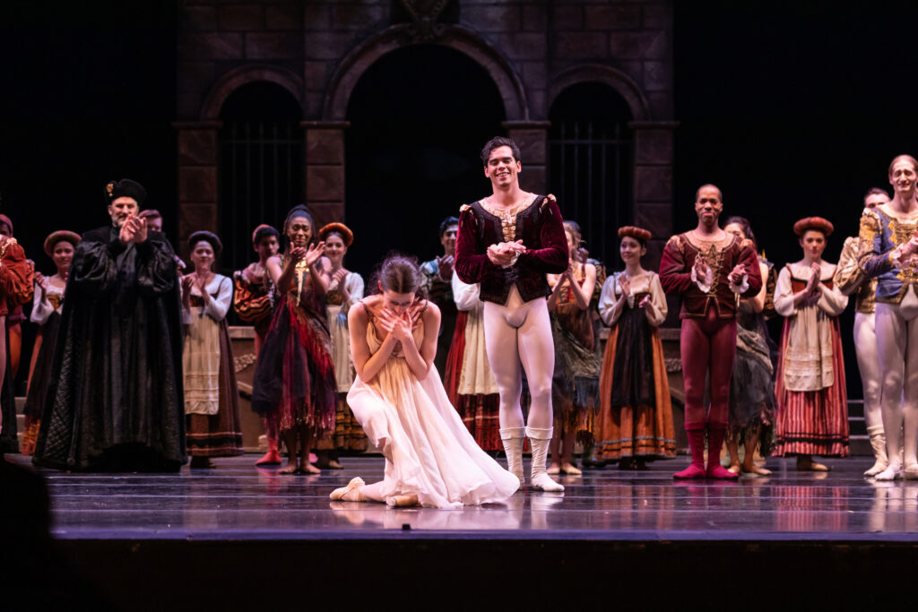 Caitlin Valentine bows deeply center stage after a performance of Romeo and Juliet. She is costumed in a filmy pink dress, tights and pointe shoes and wears her long hair pulled back away from her face. Her dance partner stands slightly behind her dressed in a red velvet tunic and white tights and boots, and claps towards her. A large group of dancers also in costume stand in the background in various Renaissance-style costumes, applauding.