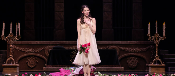 Caitlin Valentine stands center stage, her feet in a small first position, holding pink roses in her right hand and touching her chest with her left hand. She smiles gratefully toward the audience. She wears a filmy, cream colored calf-length dress with an empire waist. She wears her long dark hair pulled back near her face. Bouquets of flowers are scattered around her on the stage floor.