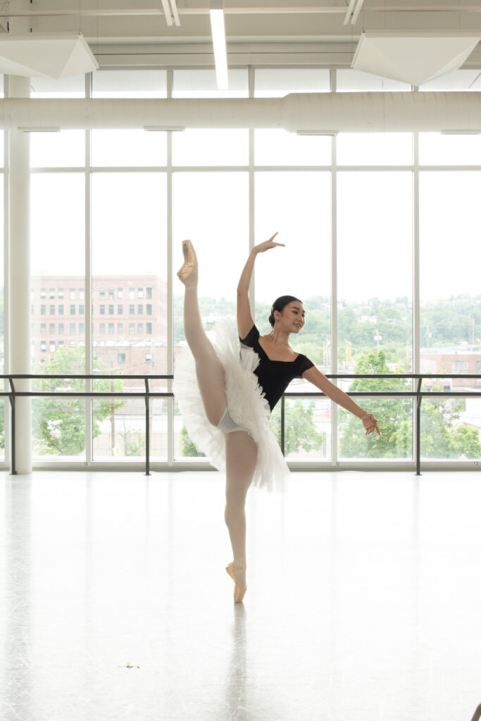 Grace Li, a teenage ballet student, does a developpé ecarté on pointe, lifting her right leg. She dances nn front of a large floor-to-ceiling window. She wears a cap-sleeved black leotard, a white practice tutu, pink tights and pink pointe shoes.