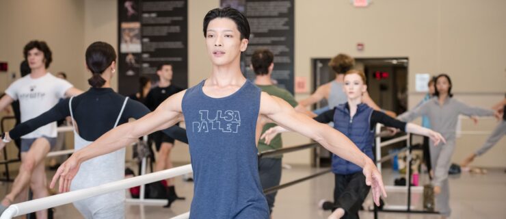 Jun Masuda takes ballet class in a large dance studio with his fellow company members. They wear various dancewear and work at portable barres in the middle of the room. Masuda is shown with his arms in second position as he balances at the barre. the other daners are in the same pose, with their left leg held 45 degrees in front of them in a degagé relevé.