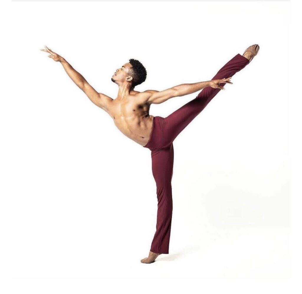 Leonard Perez performs a high first arabesque facing stage right. He is bare-chested and wears burgundy yoga pants and brown ballet slippers. He dances in front of a stark white background.