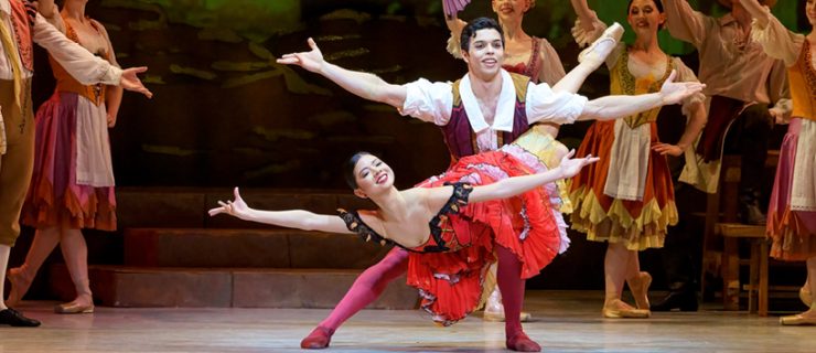 Mikaela Santos and Sergio Masero dance a pas de deux in the ballet Don Quixote. Masero lunges on his left leg, extending his right leg out slightly, and balances Santos on his left thigh as she lays across him in a fish dive, hooking her right leg around his waist and holding her upper back up. Both dancers extend their arms out triumphantly and looks towards the audience with large smiles. Santos wears a bright red, Spanish style dress and pointe shoes while Masero wears burgundy tights and shoes, a gold and burgundy vest and a white shirt. Behind them, dancers dressed as villagers watch and react happily.
