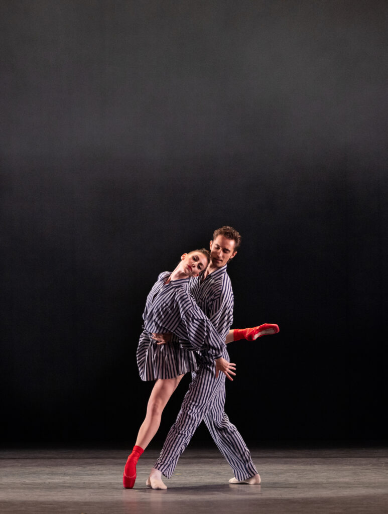 Samantha Hope Galler leans against Stanislav Olshanskyi as she does an attitude back on pointe. Olshanskyi holds her close around teh waist and pulls her slightly back. They both wear black and white striped costumes. Galler also wears red socks and red pointe shoes.