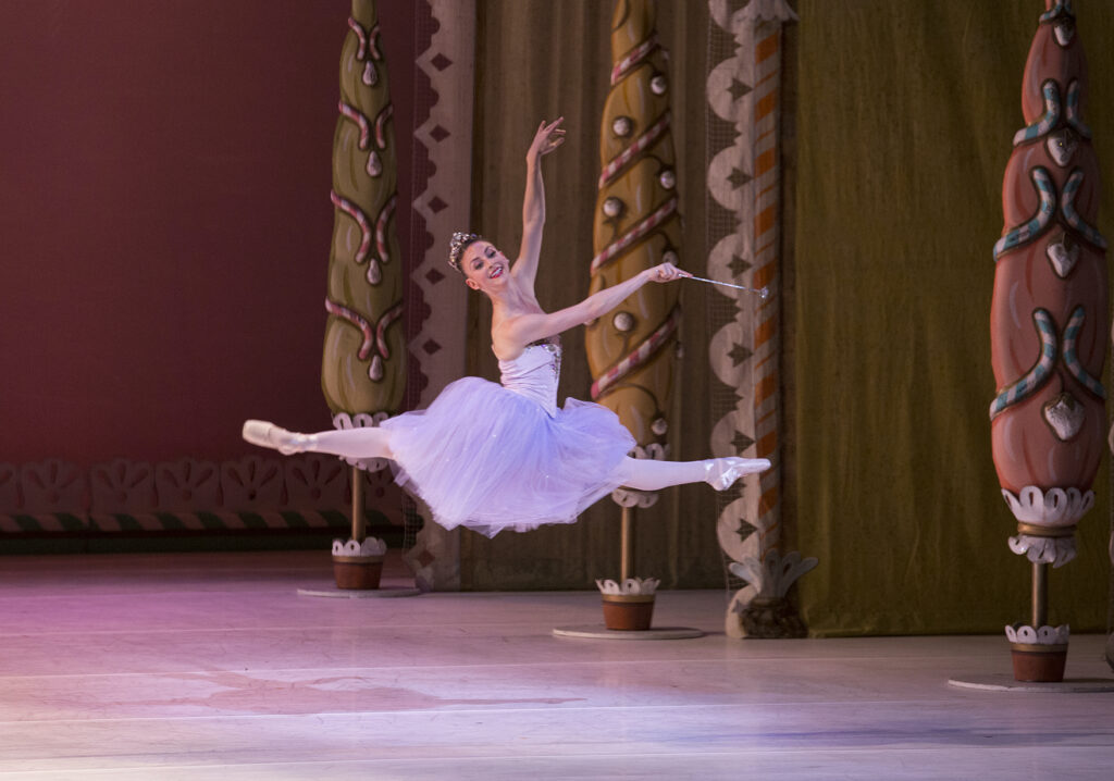 Samantha Hope Galler does a saut de chat with her arms in third arabesque towards stage left during a performance of The Nutcracker. She is costumed in a knee-length pink tutu, pink tights, pointe shoes and a sparkly tiara. She looks out towards the audience and smiles and holds a thin silver wand in her right hand.