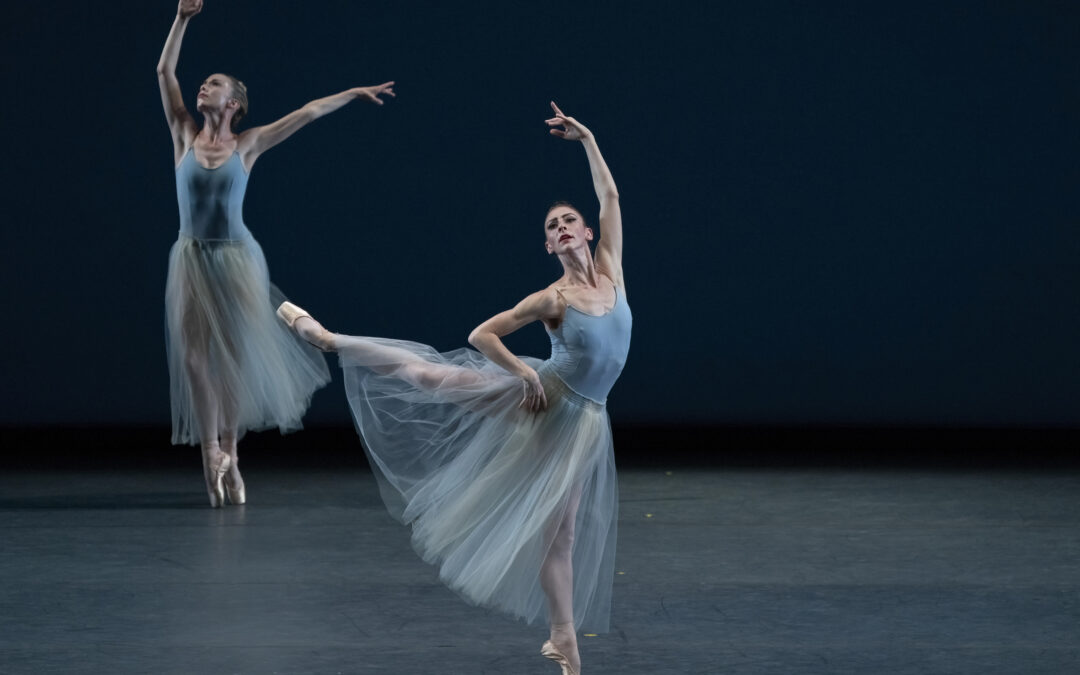 During a performance, ballerina Samantha Hope Galler, wearing a long, ankle-length blue tutu and blue bodice, does a sissone arabesque on pointe on her left leg. She holds her curved, left arm high and puts her right hand on her waist and looks out towards the audience. Another dancer in the background does a sus-sous as she moved her arms up through a position. They dance in front of a dark blue backdrop.