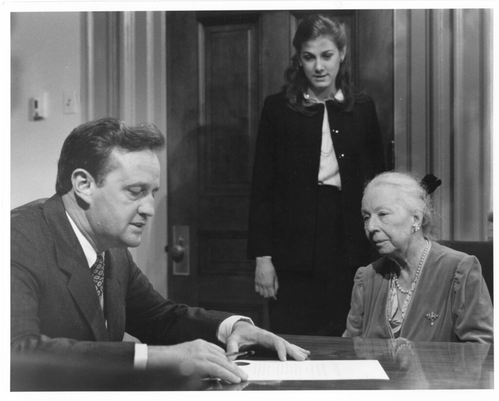 Stoner Winslett stands behind a desk at which Governor John N. Dalton and Agnes de Mille sit, discussing a piece of paper before them.