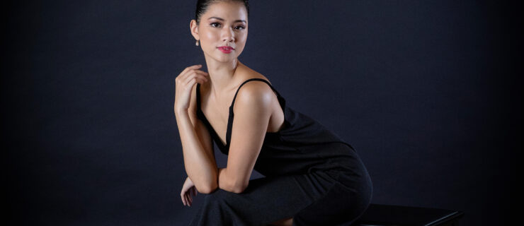 Mikaela Santos, wearing a sleek black dress, sits on a black bench in front of a black backdrop. She sits forward, propping her forearms on her knees and looks toward the camera with a small smile.