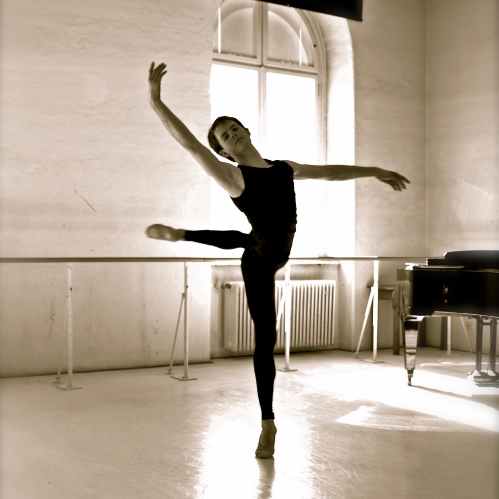 In this black and white photo, Shane Wuerthner does an attitude renversé in a sunlit dance studio. He wears a black tank top and black tights and ballet slippers. A piano is seen in the righthand corner.