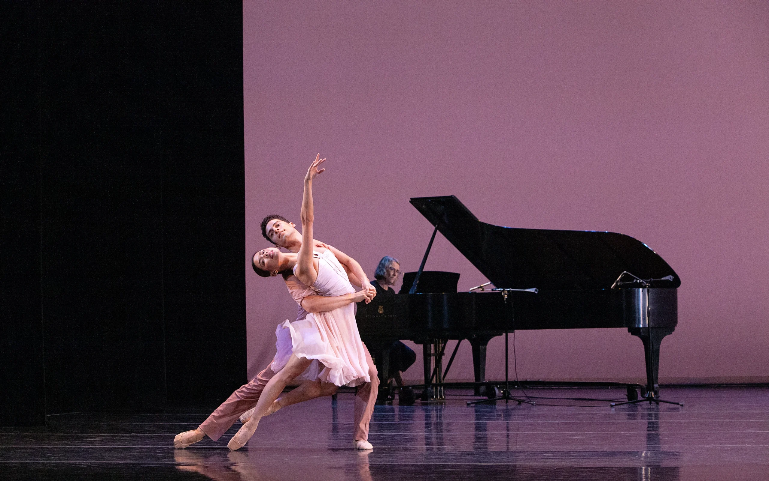 MIkaela Santos and Keaton Leier perform a sweeping pas de deux onstage. Leier, who wears mauve pants, lunges to his left, holding Santos around the waist. On pointe in a coupé back, Santos arches back and reaches her right arm up. She wears a oink chiffon dress. Behind them upstage, a woman plays a black grand piano.