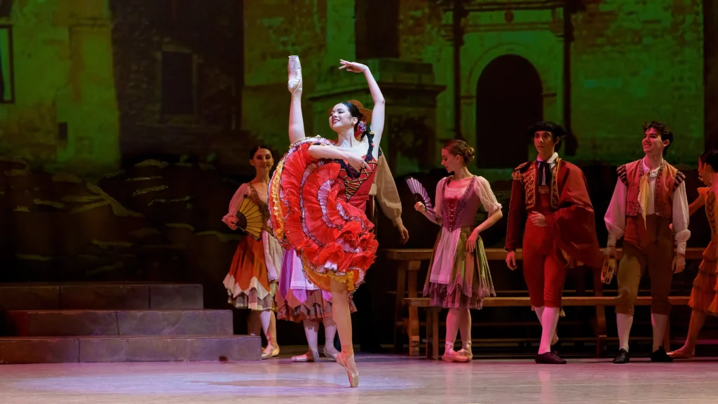 Mikaela Sanotos does a high grand battement with her right leg during a performance of Don Quixote. She wears a red Spanish-style dress with a ruffly skirt and red flowers in her hair, and she smiles triumphantly as she looks toward her lifted leg. Behind her, male and female dancers in Spanish-style costumes watch and smile..