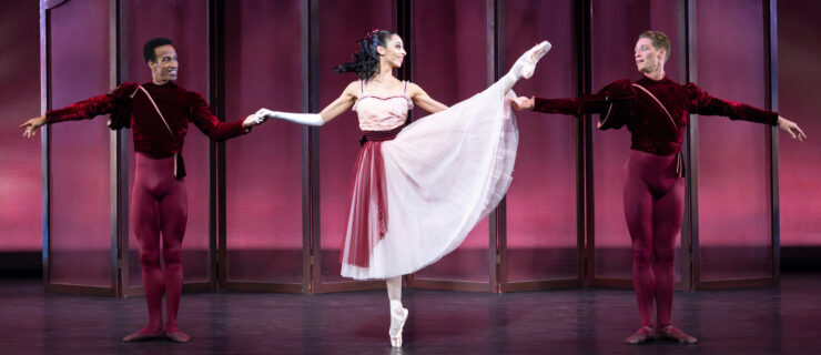 Ricardo Rhodes, Jessica Assef, and Daniel Pratt, three professional ballet dancers, perform a pas de trois onstage in front of a burgundy backdrop. Assef, wearing a light pink calf-length tutu with a burgundy sash, stands between the men, raises her left leg in développé a la seconde on pointe. She holds her arms out to the side, holding each partner's hand, and looks toward Pratt on her left. The men, wearing burgundy tights and matching velvet tunics, stand in a small first position and smile at Assef.