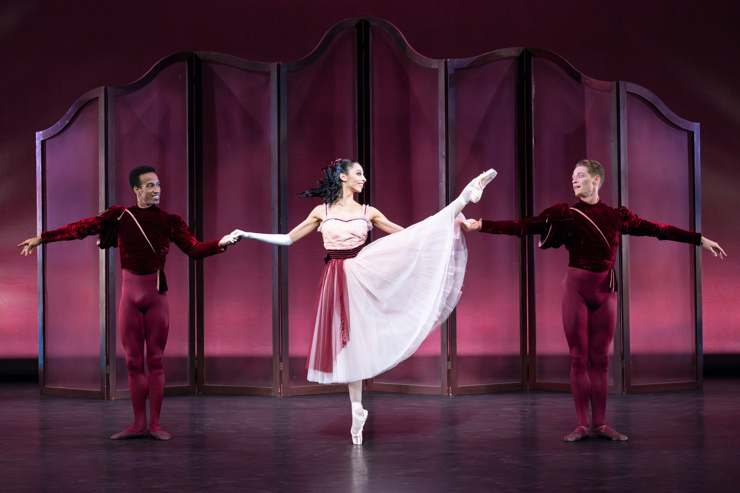 Ricardo Rhodes, Jessica Assef, and Daniel Pratt, three professional ballet dancers, perform a pas de trois onstage in front of a burgundy backdrop. Assef, wearing a light pink calf-length tutu with a burgundy sash, stands between the men, raises her left leg in développé a la seconde on pointe. She holds her arms out to the side, holding each partner's hand, and looks toward Pratt on her left. The men, wearing burgundy tights and matching velvet tunics, stand in a small first position and smile at Assef.