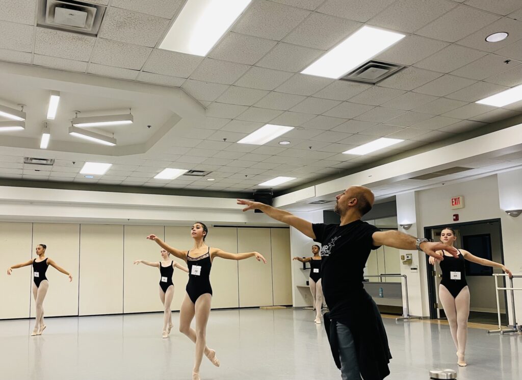 Roberto Forleo teaches ballet class in a dance studio. He is shown in profile, reaching his arms out in first arabesque. He wears jeans and a black t-shirt. Behind him, a group of five female dancers do the same arm position and do a step on relevé. They wear black leotards, pink tights, and pink ballet slippers. They all have a number pinned to the front of their leotards.