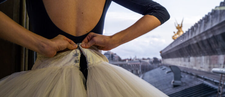 A close-up shot of a young female dancer from the back shows her fastening her white practice tutu, with the Palais Garnier roof in the background.