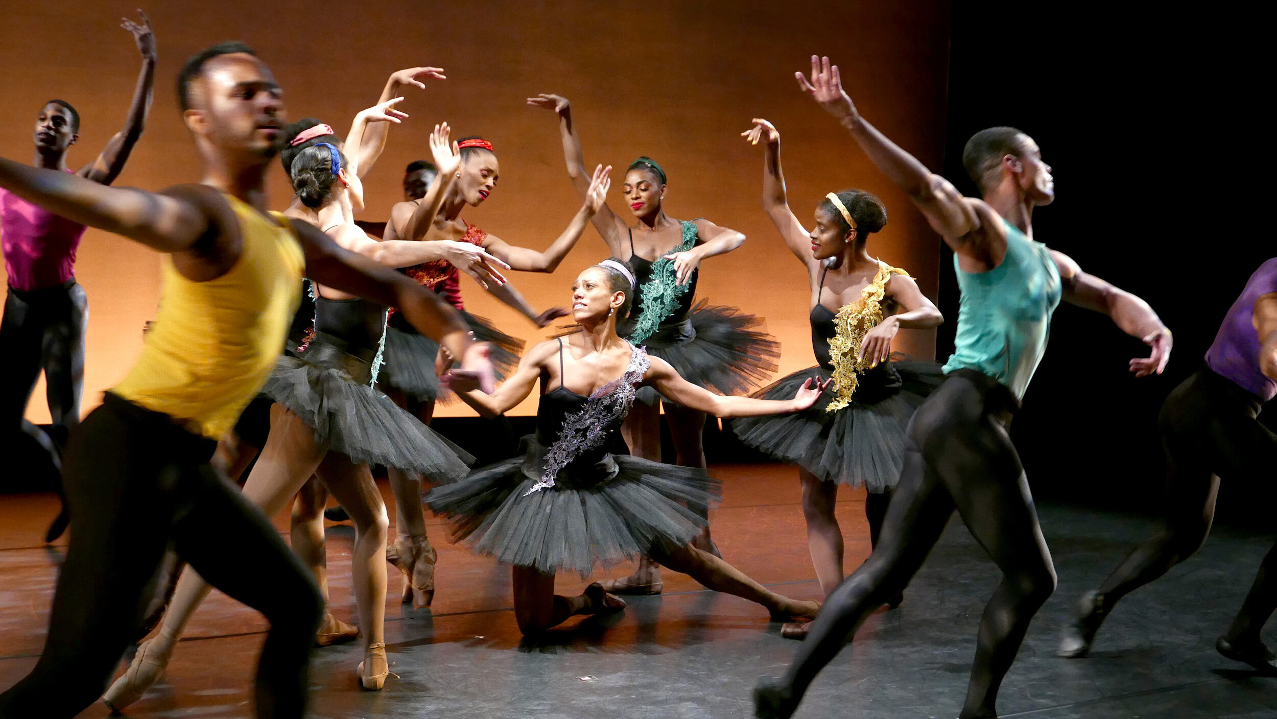A ballerina in a black tutu kneels onstage onto her right knee and opens her arms, looking up at a group of other female ballet dancers who surround her in various poses. A group of male dancers in black tights and brightly colored sleeveless t-shirts run in a large circle around them.