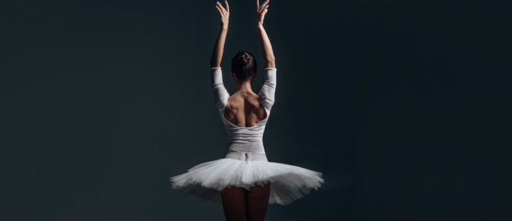 A female ballet dancer, shown with her back to the camera, poses with her arms up in a swan port de bras position. She wears a long-sleeved white leotard and a white pancake tutu.