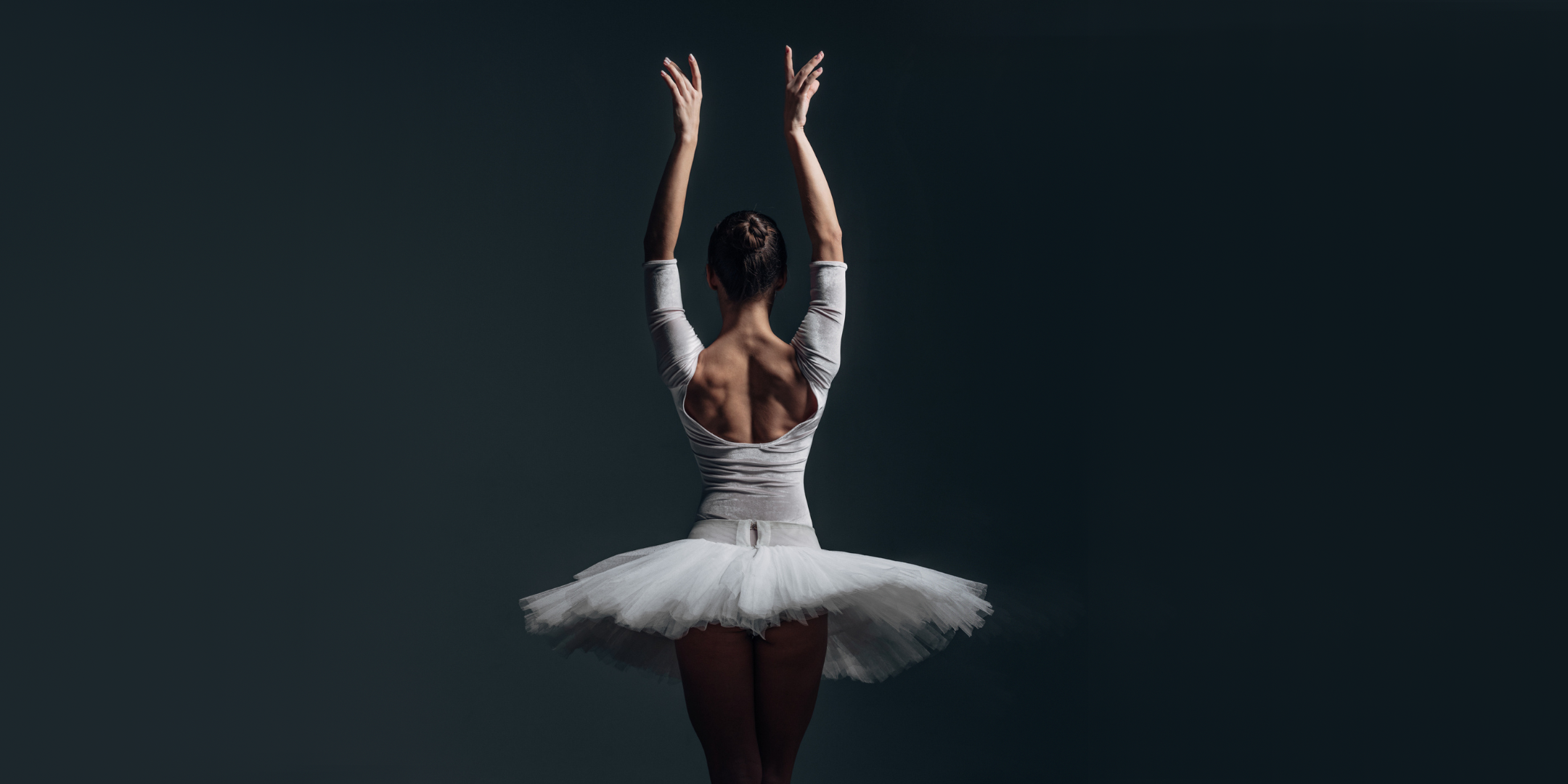 A female ballet dancer, shown with her back to the camera, poses with her arms up in a swan port de bras position. She wears a long-sleeved white leotard and a white pancake tutu.