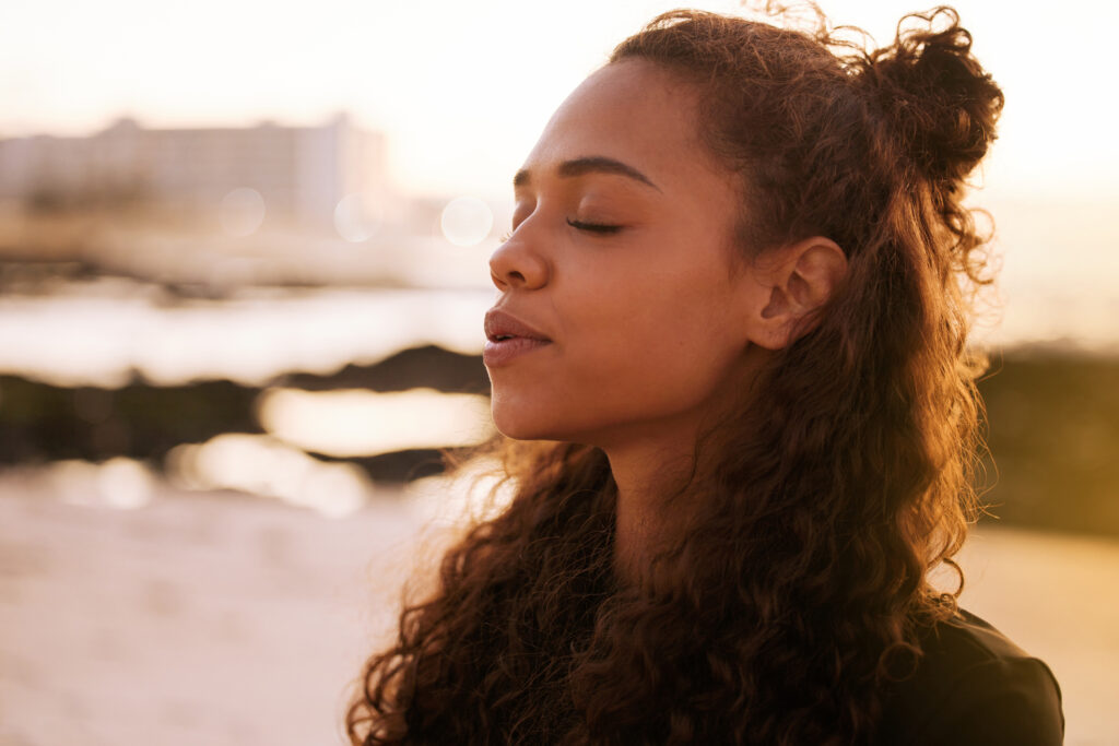 A close-up photo of a young woman sitting alone on a mat and meditating on the beach at sunset