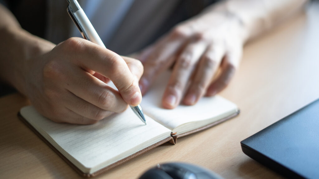 A close-up photo of a man's hands shows him writing in a notebook