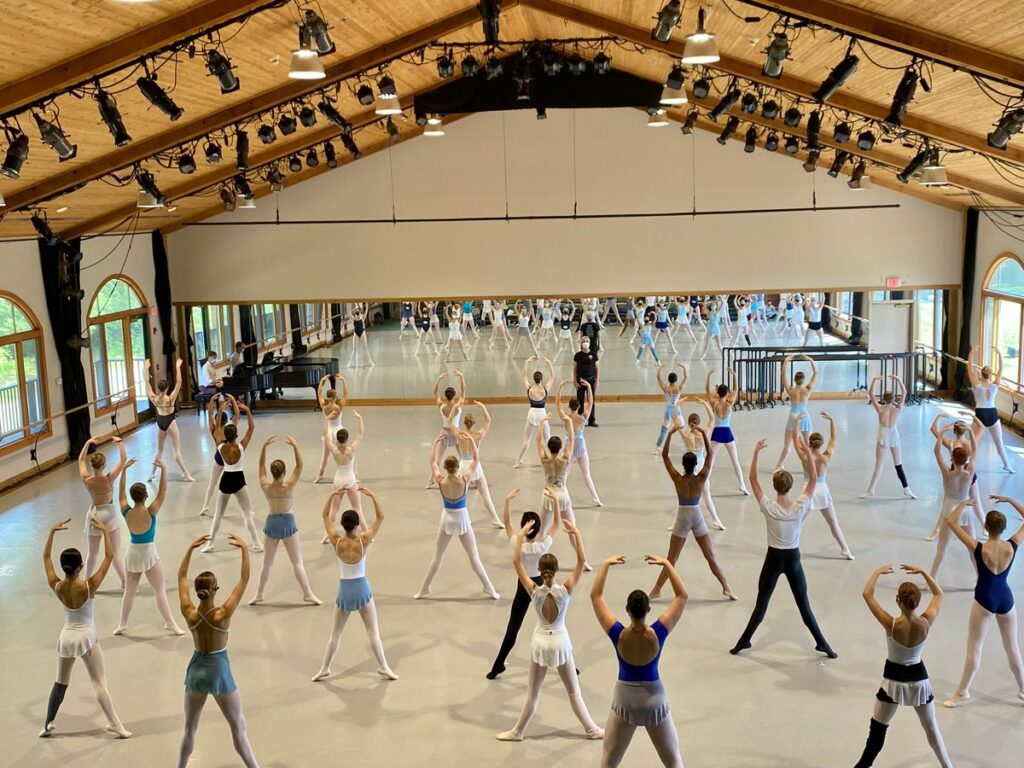 A large class of ballet dancers is shown from behind in a large dance studio with vaulted ceilings. They face the mirror and stand in second position relevé with their arms in high fifth. A male teacher stands at the front of the room, facing them, and an accompanist plays a piano in the front corner.