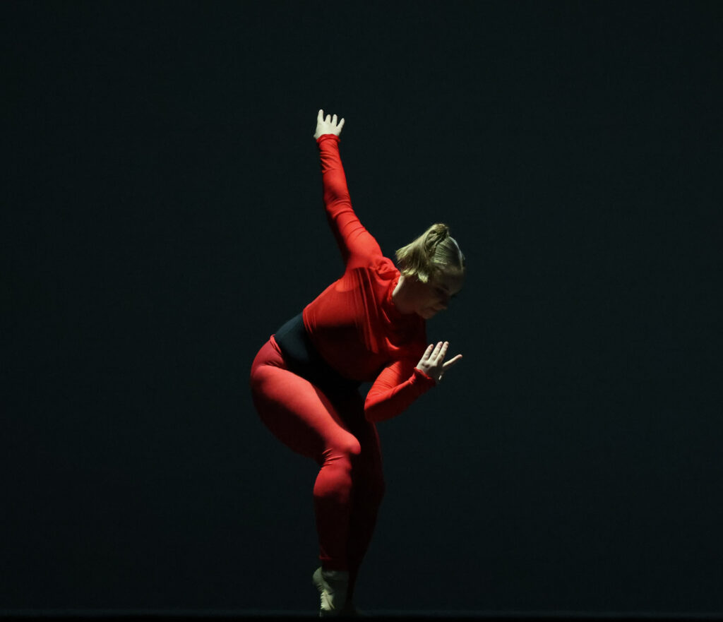 Kirsten SMerus is shonw dancing on a dark stage within a bright spotlight. She crouches down slightly, reaching her right arm up. She wears red tights with a black leotard underneath and a sheer, cropped long-sleeved shirt.