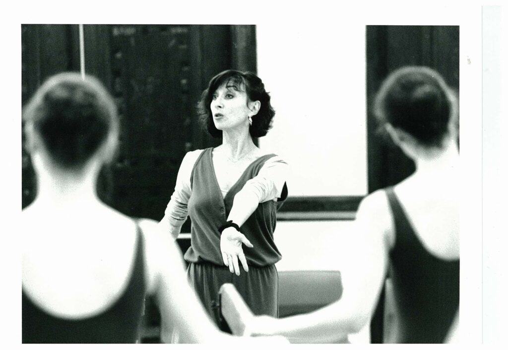 IN this black and white photo, Mavis Staines is shown teaching a ballet class, exteneding her left arm out in front of her and flexing her wrist. She wears a dark, v-neck jumper over a long-sleeved shirt. In the foreground we see the backs of two female ballet students in dark tank leotards, who stand at a barre and listen.