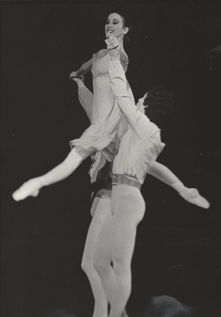 In this black and white photo, two male dancers lift Mavis Staines high into the air, holding her armpits and forearms. Staines weara a knee-length, empire-waisted dress with a flowing chiffon skirt. Her legs are spit slightly and she looks out with a smile.