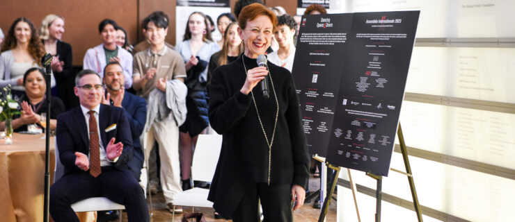 Mavis Staines, wearing a black sweater and black pants, stands next to an easel with two presentation posters and speaks into a microphone, smiling broadly. A crowd of people is shown sitting and standing behind her, applauding.