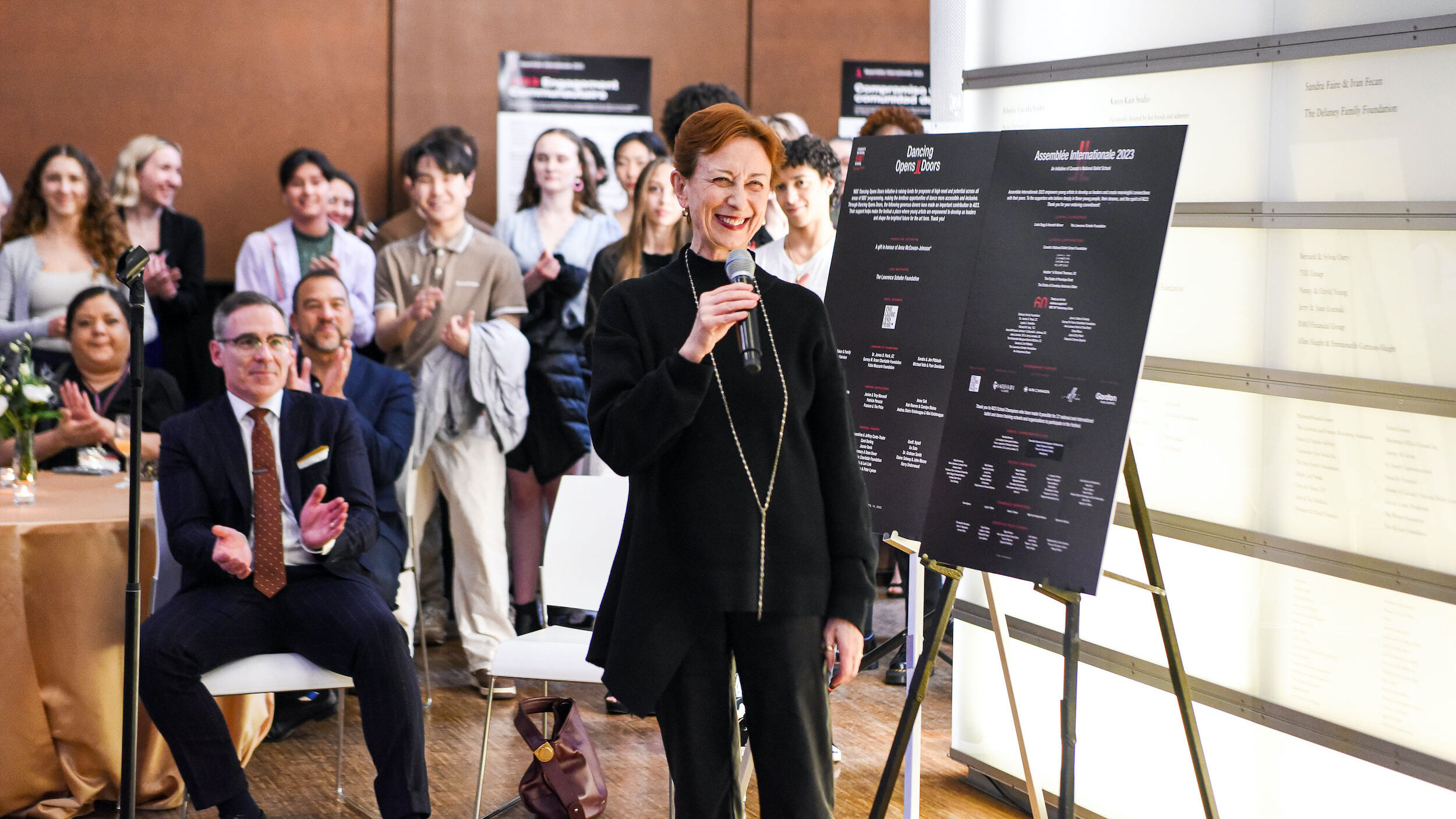 Mavis Staines, wearing a black sweater and black pants, stands next to an easel with two presentation posters and speaks into a microphone, smiling broadly. A crowd of people is shown sitting and standing behind her, applauding.