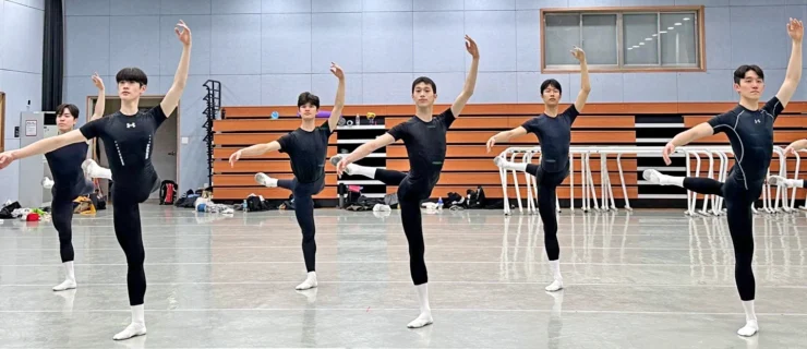 In a large ballet studio, six male ballet students in their late teens and early 20s are shown in two staggered lines practicing attitude derriere croisé with their left leg up. They all wear black t-shirts and tights, white socks and white ballet slippers.