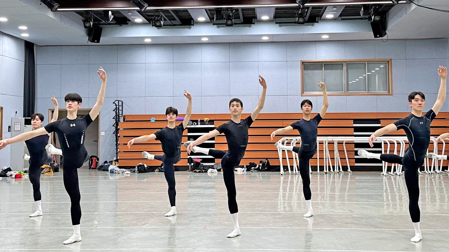 In a large ballet studio, six male ballet students in their late teens and early 20s are shown in two staggered lines practicing attitude derriere croisé with their left leg up. They all wear black t-shirts and tights, white socks and white ballet slippers.