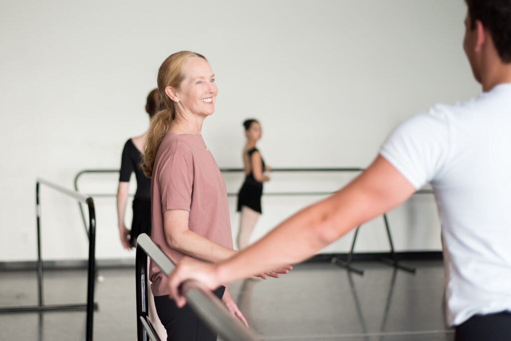 Margaret Tracey stands with her back at the barre wearing a pink 3/4 sleeve shirt and black pants. She smiles as she speaks to her ballet class.