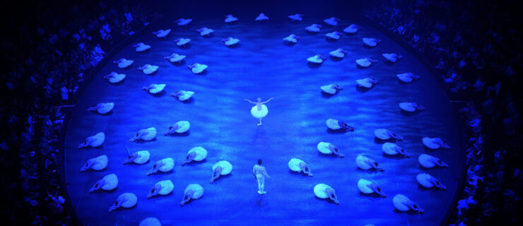 A circular stage is shown from above, lit in blue, during a performance of Swan Lake. A large corps de ballet in white tutus sits on the ground with one leg extended in front of them, their upper bodies bent over their legs. They make a circular formation. In the middle, a ballerina in a white tutu and feathered crown stands in tendu derriere and extends her arms out to her sides. She faces a male danseur, costumed in a white tunic and tights. The audience surrounds them on all sides.