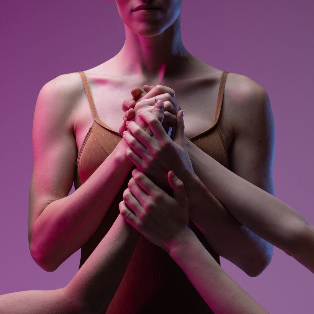 A close-up of a female dancer's torso, cut off just above the tip of her nose. She holds her hands in front of her chest, clasping on to several pairs of hands as other arms extend into the photo.