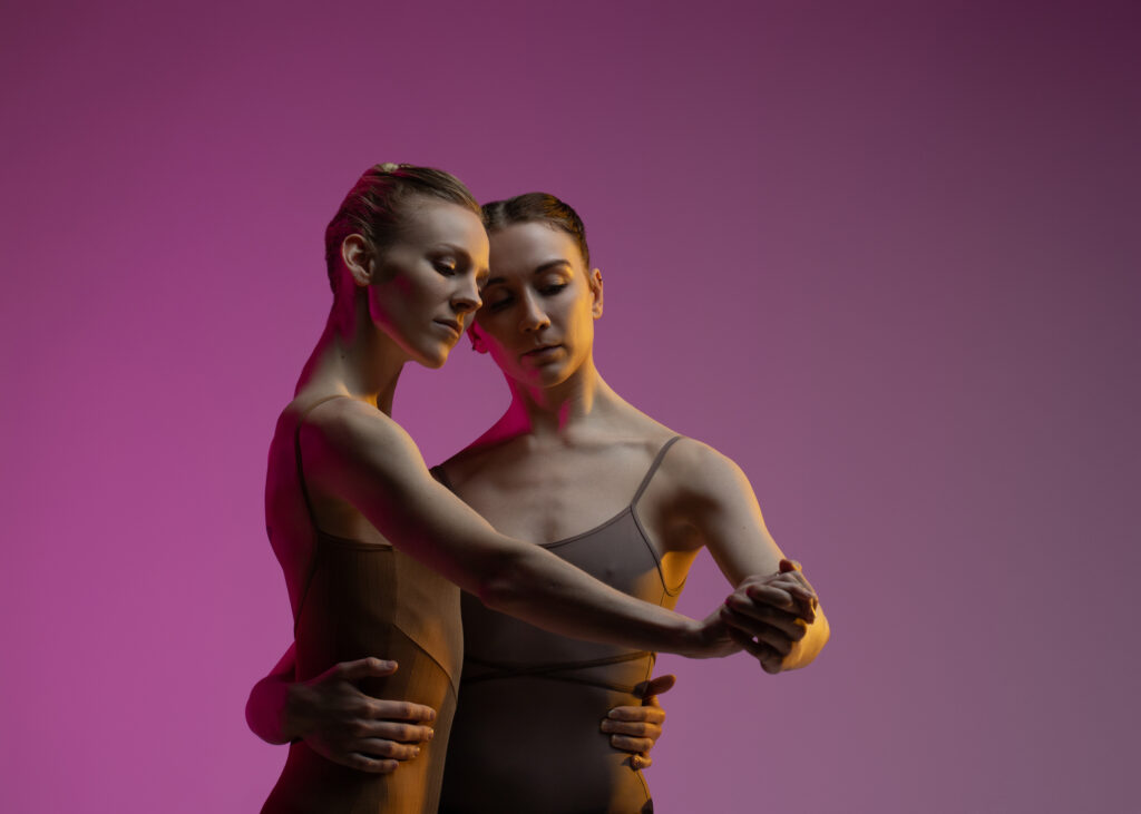 Demi Trezona and Sara Jumper, shown hips-up, hold hands and wrap their arms around each others' waists in a waltz posture.
