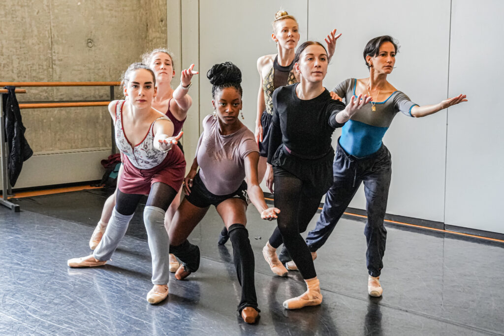 The cast of <i>Dream of a Common Language</i> rehearses: six female dancers lunging forward together in a clump, left arms outstretched as if asking or calling to action.